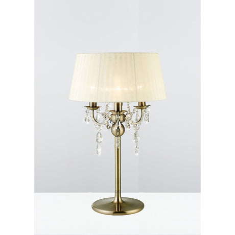 The Olivia 3 Light Crystal Table Lamp in antique brass features a round base, cream pleated shade, and crystal accents on curved arms, shown against a plain light background.