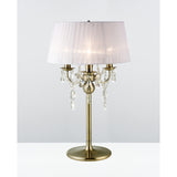 The Olivia 3 Light Crystal Table Lamp in antique brass features a white pleated shade, three candle-style bulbs, and hanging crystal accents, adding elegant lighting to any space. Displayed on a white surface with a plain light background.