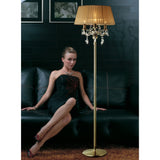 A woman in a black dress with a red web design sits on a leather sofa. In the foreground stands the Olivia 3 Light Crystal Floor Lamp in brass with a bronze shade. The stylish scene is set by dark walls and framed pictures.