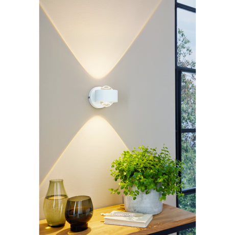 Ono 2 LED Wall Light in white aluminium mounted on a wall, casting warm up and down light, shown above a wooden console with a plant and vases.