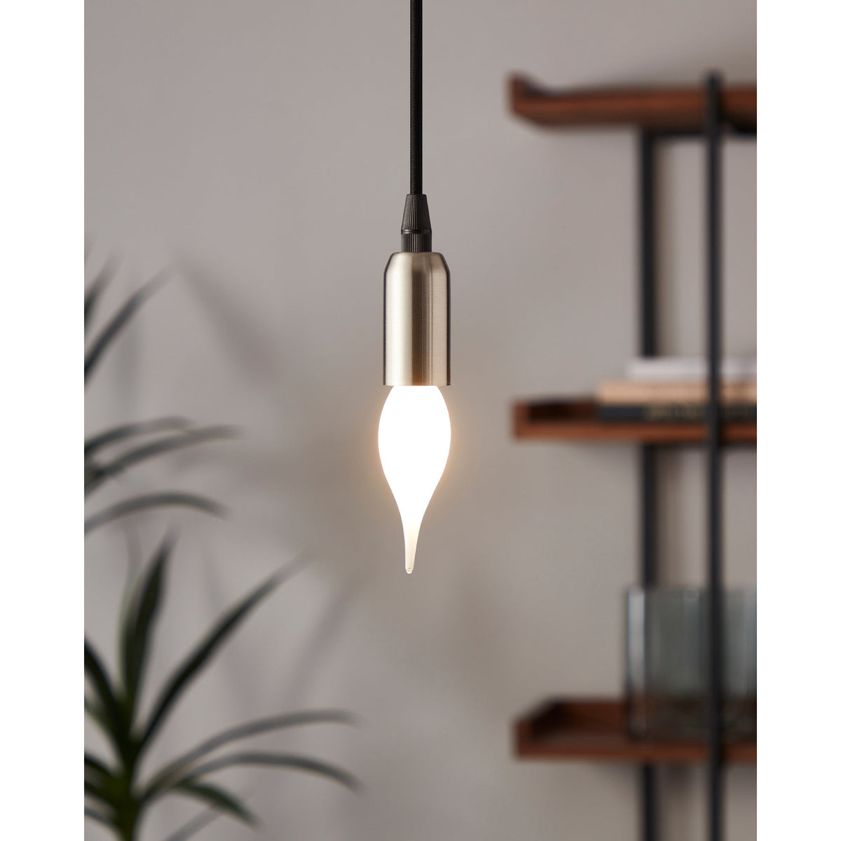 Opal LED bulb with flame-tip candle shape in a brushed metal pendant light, glowing warm white against a neutral living room backdrop with shelving and a plant.