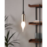 Opal LED bulb with flame-tip candle shape in a brushed metal pendant light, glowing warm white against a neutral living room backdrop with shelving and a plant.