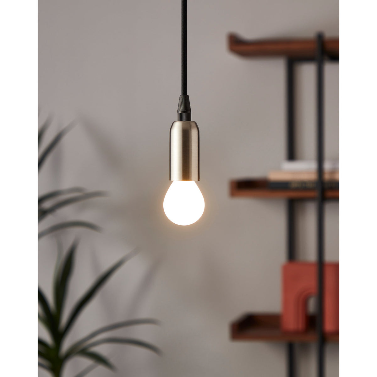 Opal LED golf ball bulb glowing warm white in a brushed metal pendant light fitting, shown in a room setting with a plant and shelving in the background.