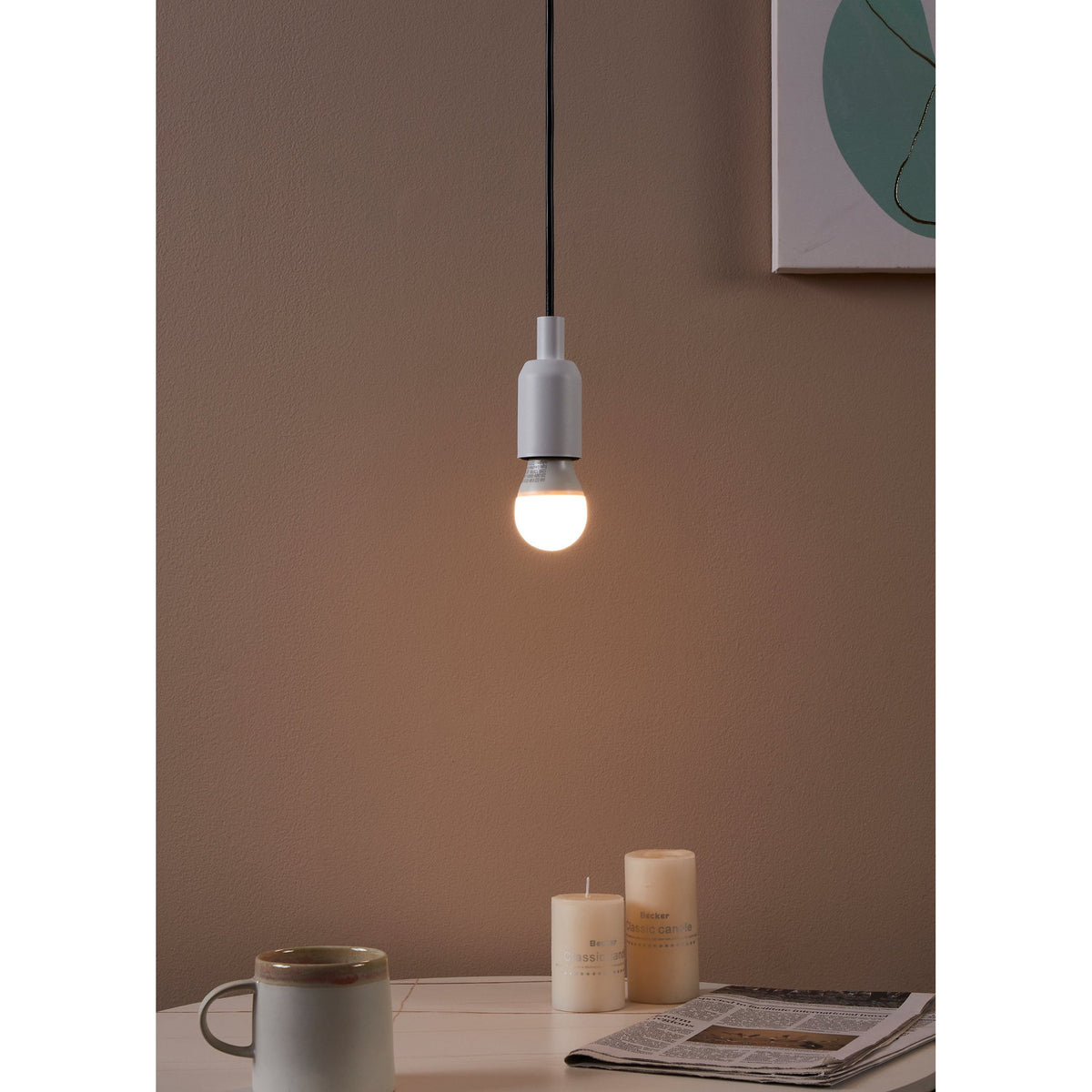 Opal LED golf ball bulb, E27 fitting, glowing warm white, hung from a grey pendant holder above a table with candles and a mug in a muted room setting.