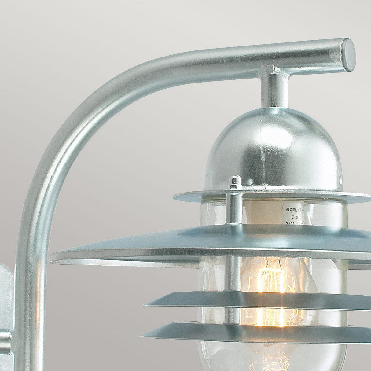 The Oslo Outdoor 1 Light Wall Lantern - Silver is presented in a close-up, highlighting its metallic industrial style with a distinctive curved arm design. Made from galvanized steel, this IP55-rated fixture includes layered metal shades and clear glass, casting a warm glow against a smooth gray background.