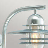 The Oslo Outdoor 1 Light Wall Lantern - Silver is presented in a close-up, highlighting its metallic industrial style with a distinctive curved arm design. Made from galvanized steel, this IP55-rated fixture includes layered metal shades and clear glass, casting a warm glow against a smooth gray background.