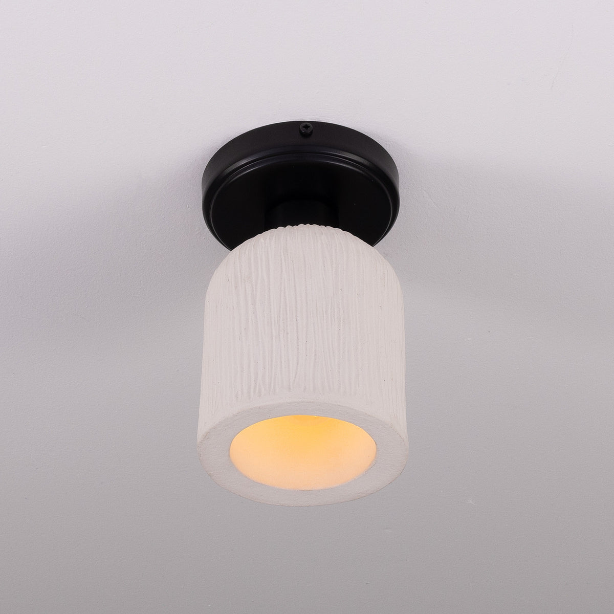 Osier Ceramic Flush Ceiling Light with a matt white striped cylindrical ceramic shade on a round black ceiling plate, warm bulb glow visible, plain light grey background.