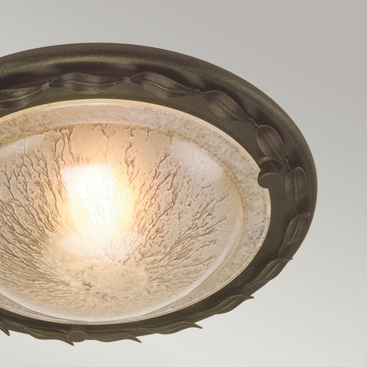 The Olivia 1 Light Flush in Black/Gold Patina emits a gentle glow through its textured, frosted glass shade. It is adorned with a decorative dark metallic rim featuring an elegant leaf-like pattern enhanced by black/gold patina. The backdrop is muted gray, adding to its sophisticated design.