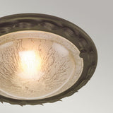 The Olivia 1 Light Flush in Black/Gold Patina emits a gentle glow through its textured, frosted glass shade. It is adorned with a decorative dark metallic rim featuring an elegant leaf-like pattern enhanced by black/gold patina. The backdrop is muted gray, adding to its sophisticated design.