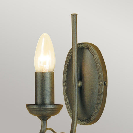 The Olivia 1 Light Wall Light - Black/Gold Patina features a vintage-style handmade design with a lit bulb resting on an ornate metal base. Mounted on a light gray wall, the bulb resembles a candle flame, enhancing the decor with its classic appeal.