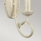 The Olivia 1 Light Wall Light - Ivory/Gold Patina is a vintage-style sconce with an elegant curved arm and raised decorative elements on the backplate, showcasing a distressed white finish. This handmade wall light features an exquisite Ivory/Gold Patina beautifully set against a gray background.