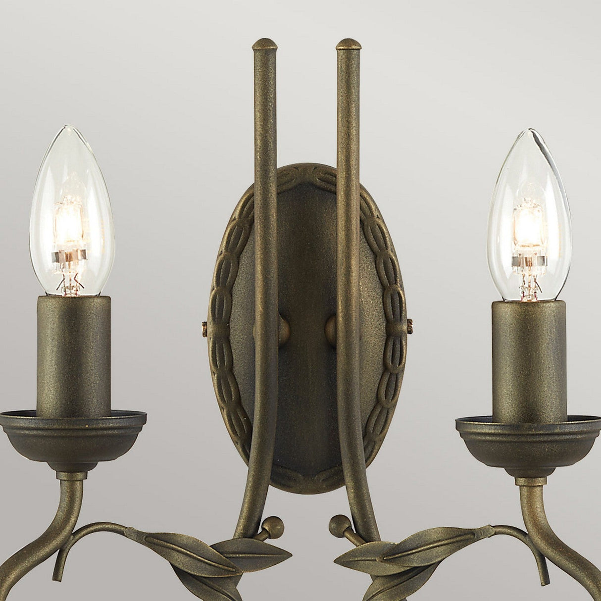 The Olivia 2 Light Wall Light - Black/Gold Patina features a rustic bronze finish enhanced by a subtle black/gold patina. Its intricate backplate and decorative leaf details complement the handcrafted design, while elegantly curved arms support candle-style bulbs, offering a vintage and classic appeal against a neutral backdrop.