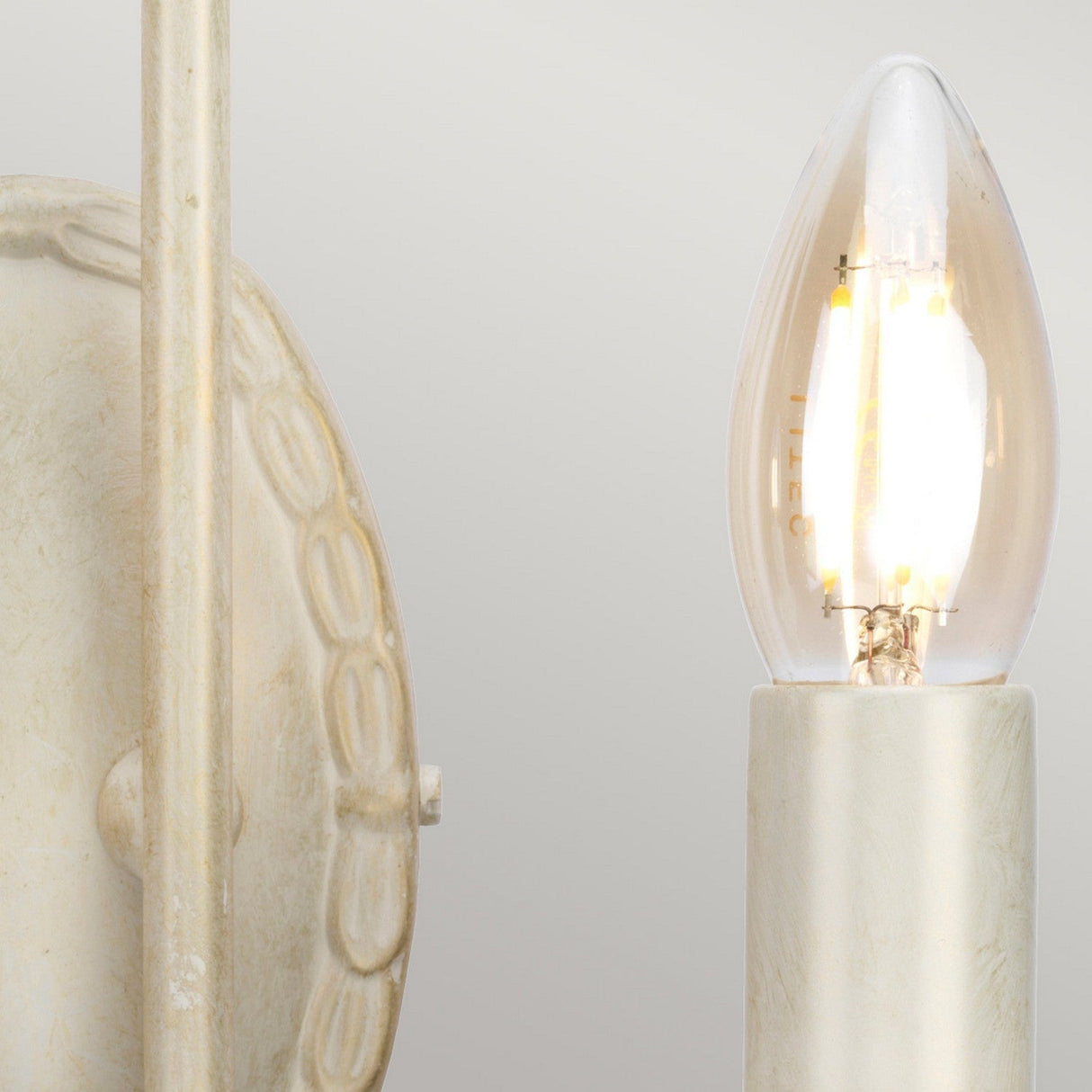 Close-up of a lit LED bulb in the Olivia 2 Light Wall Light fixture, featuring a textured, circular wall mount with an elegant ivory and gold patina. The handmade lighting fixture emits a soft, warm glow against a neutral background.