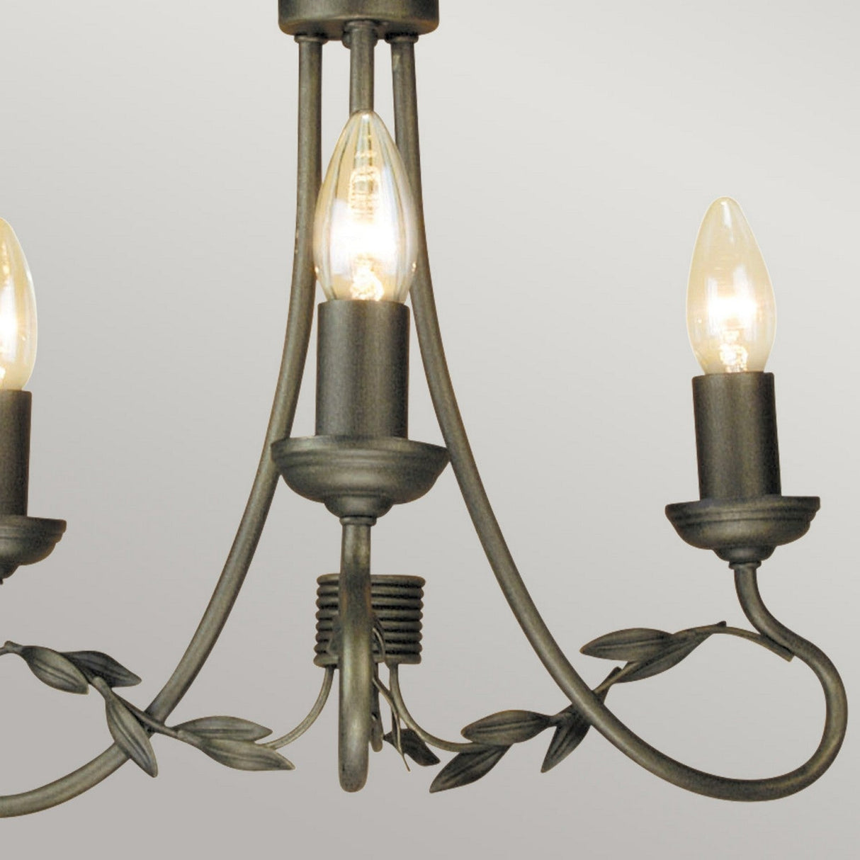 Close-up of the Olivia 3 Light Chandelier in a black/gold patina finish, featuring three candle-shaped light bulbs. Its design showcases elegantly curved arms adorned with leaf accents, all set against a plain gray background that emphasizes its graceful charm.