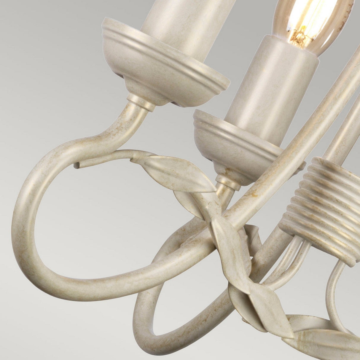 Close-up of the Olivia 3 Light Chandelier showcasing its ornate, vintage-style design with a twisted cream-colored metal structure adorned with leaf accents and an exposed bulb. This handmade piece is finished in an exquisite ivory/gold patina and appears against a backdrop of light gray tones.