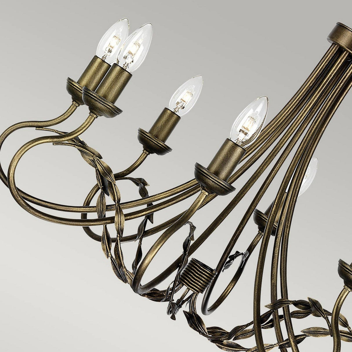 The Olivia 8 Light Chandelier in a black/gold patina is an elegant piece with intricate, swirling metalwork. It features eight candle-shaped bulbs attached to ornate, curved arms, with clear bulbs showcasing filament details against a soft gray background.