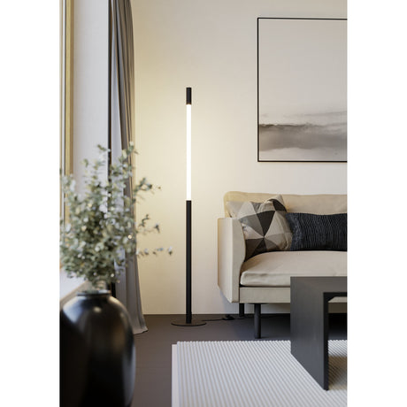 Parabita LED Floor Lamp in black and white, slim steel pole with vertical white diffuser strip on a round flat base, shown beside a sofa in a living room.
