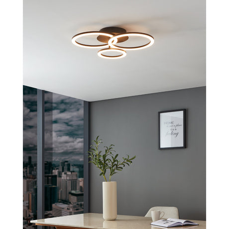 Parrapos Smart LED Flush Ceiling Light in black steel with three overlapping illuminated rings, shown mounted on a white ceiling above a dining area.