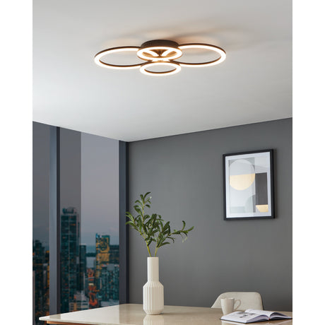 Parrapos Smart LED Flush Ceiling Light in black steel, four interlocking rings with warm white illumination, shown mounted on a white ceiling in a grey-toned dining room.