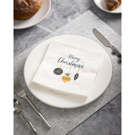 White paper napkin folded on a plate, printed with "Merry Christmas" in black script and black and gold bauble designs, set beside cutlery with bread rolls in the background.