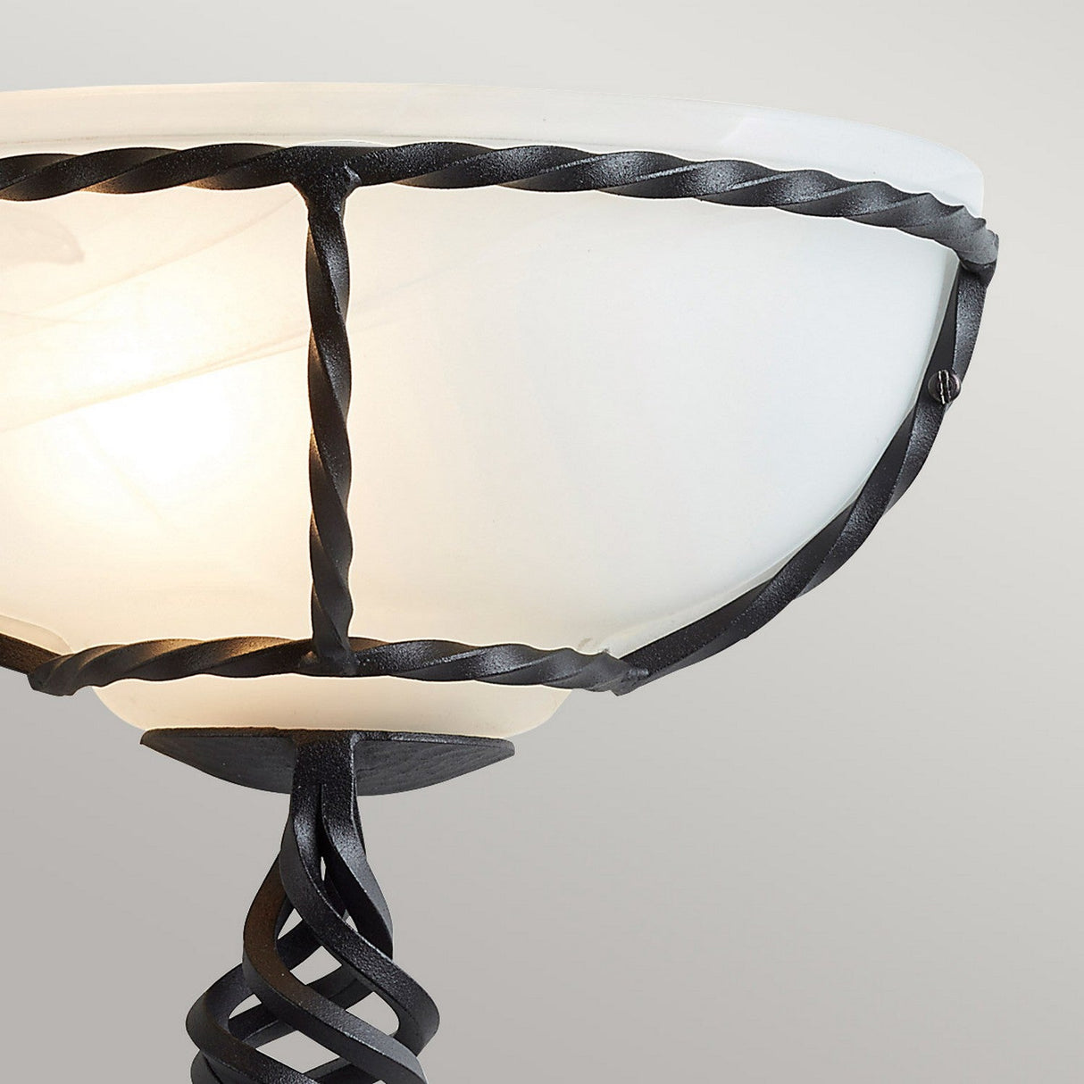 A close-up view of the Pembroke Wall Light - Black showcases its black hand-forged iron intricately twisted around a white frosted glass lampshade. The iron adorns the base and rim of the bowl-shaped glass, which emits a gentle glow, making it stand out against the plain off-white background.