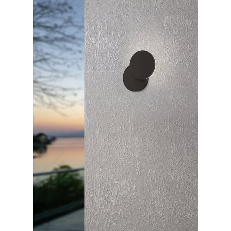 Pedersano round LED outdoor wall light in black aluminium and plastic, mounted on a textured grey wall, casting a soft edge glow, with a blurred dusk waterside view alongside.
