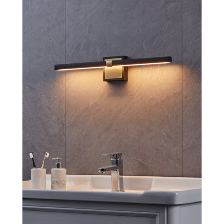 Peguera 60cm LED bathroom wall light in black and brushed brass, slim horizontal bar fixture mounted above a bathroom sink with warm white glow, set against a grey stone-effect wall.