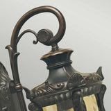 Close-up of the Philadelphia 1 Light Outdoor Medium Down Wall Light, featuring an ornate vintage-style design with a curved handle and intricate metalwork. This wall light is finished in old bronze, showcasing decorative patterns and a glass panel against a light, plain background.