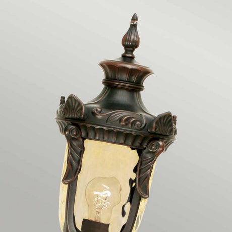 The Philadelphia 1 Light Medium Pillar - Old Bronze is an ornate vintage-style lantern featuring a decorative old bronze finish and an exposed bulb inside. Ideal for outdoor lighting, it showcases intricate scrollwork and leaf motifs on a gray background.