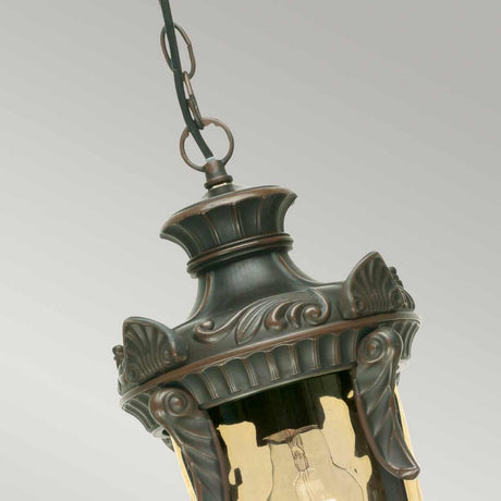 Close-up of the Philadelphia 1 Light Outdoor Medium Chain Pendant in old bronze. The metal casing showcases intricate scrollwork and decorative elements, while a clear glass shade reveals the bulb inside. This vintage-style outdoor chain pendant is suspended against a plain background.