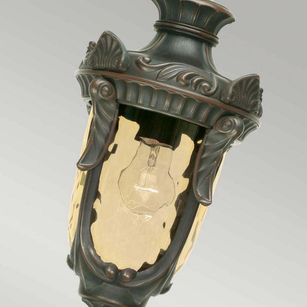 Close-up of the Philadelphia 1 Light Outdoor Medium Chain Pendant, featuring a clear glass cover with a visible bulb inside. The pendant is suspended outdoors and showcases an old bronze metal frame adorned with intricate designs that exude period-style elegance. The backdrop is a soft gray.