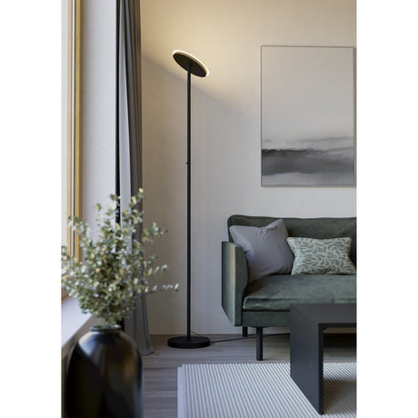 Piagnolo LED Floor Lamp in black, with a slim steel stem, round base, and flat round head, shown in a living room beside a green sofa and framed artwork.