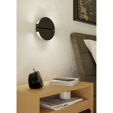 Pianosa LED Wall Light in black, round split-disc design with slim horizontal slot, mounted above a bedside table in a bedroom setting.