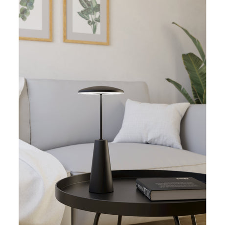Piccola LED Touch Table Lamp in black, with a flat mushroom-shaped shade on a slim steel stem and tapering base, shown lit on a side table in a living room setting.