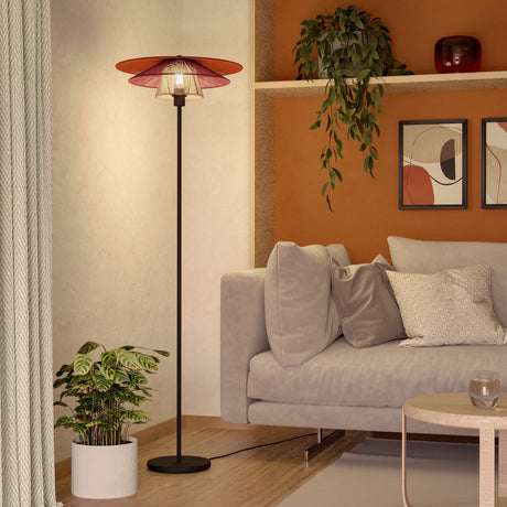 Pignona Floor Lamp in black steel with a tiered terracotta, burgundy and cream textile shade, shown on a round base in a living room beside a sofa and plants.