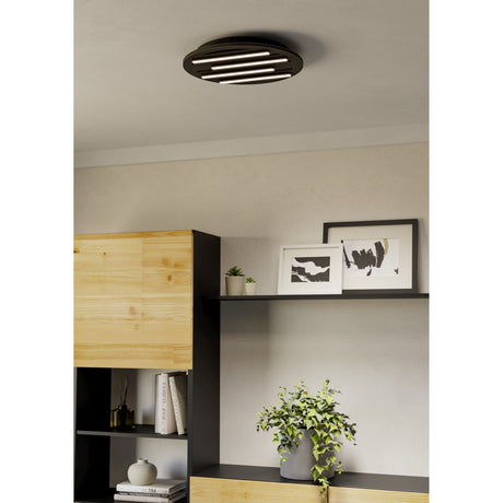 Pitrignani round LED flush ceiling light in matt black, 38cm, with slatted face and staggered LED strips, shown installed on a living room ceiling above a shelving display.