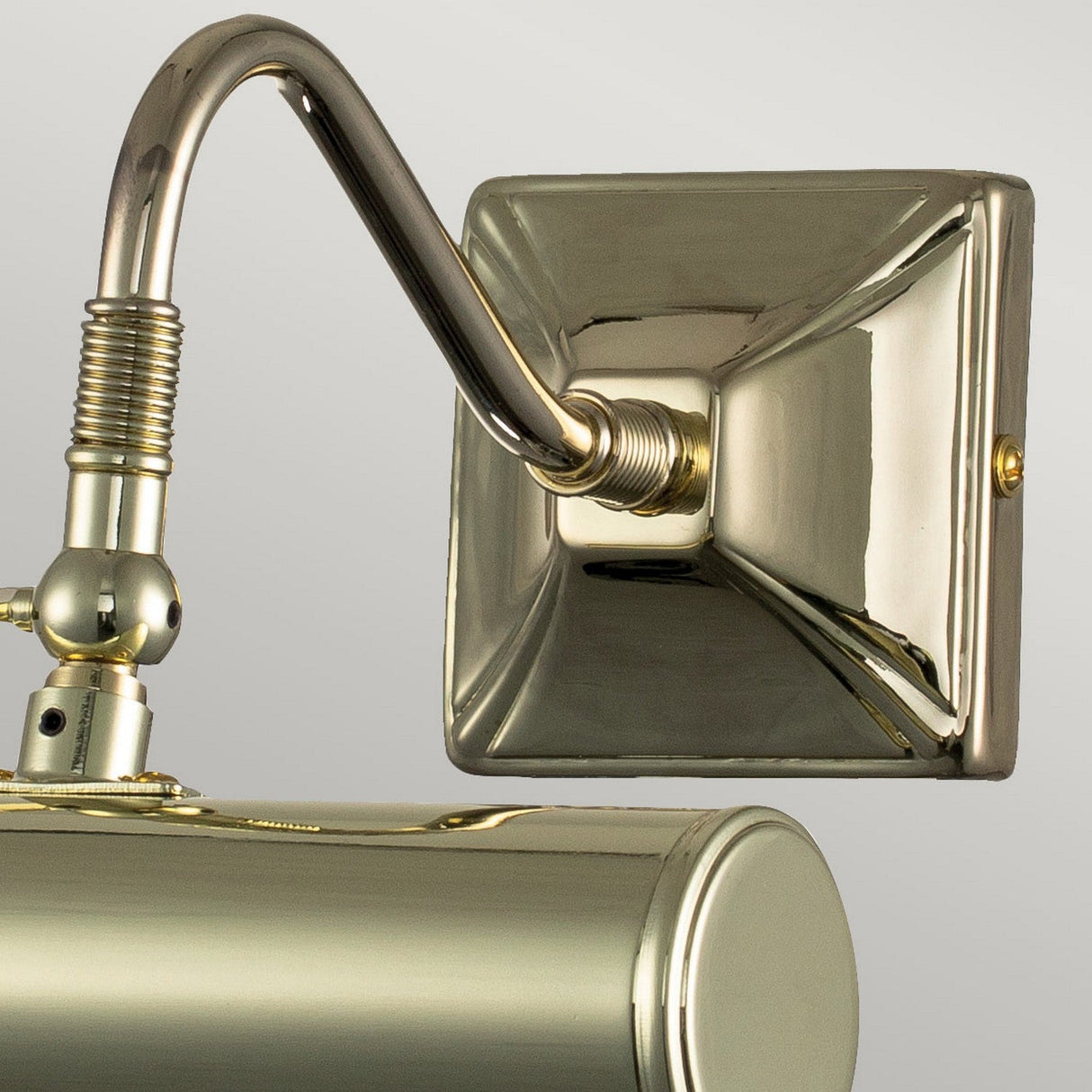 A close-up of the Picture Light 1 Light Small in polished brass showcases its shiny, rectangular base and elegantly curved arm. The metallic surface beautifully reflects light, highlighting its smooth and polished finish against a plain gray background, making it perfect for illuminating artwork.