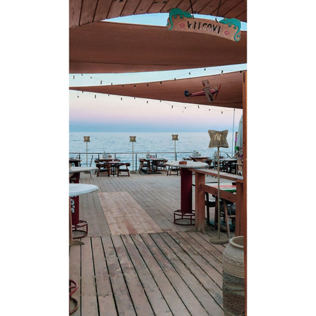 Open-air wooden deck restaurant by the sea, with tables under shade sails and string lights. A “Welcome” sign greets guests, while Polinesia 1 Light Outdoor Floor Lamps in beige add cozy garden lighting as the calm ocean shimmers nearby.