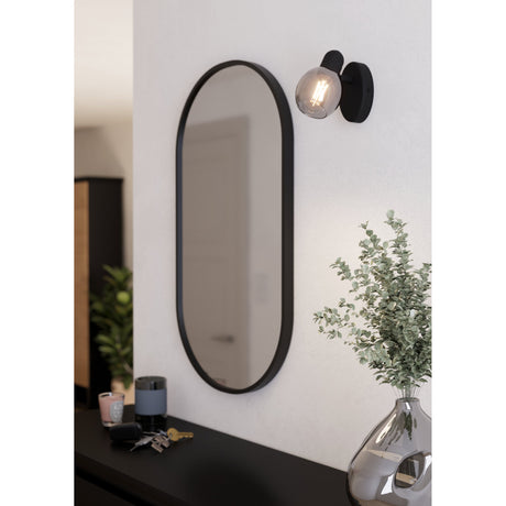 Pollica Wall Spotlight in black with round steel wall plate, short arm, and smoke-tinted glass globe, shown mounted beside an oval black-framed mirror in a hallway.