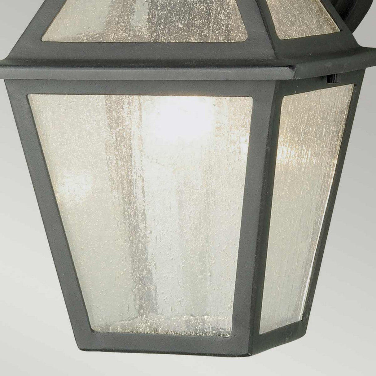 Close-up of the Polruan 1 Light Outdoor Wall Light in black, showcasing its IP44 rating and seeded glass panels encased in a dark metal frame. The fixture casts a warm glow through the textured glass, creating a cozy ambiance.