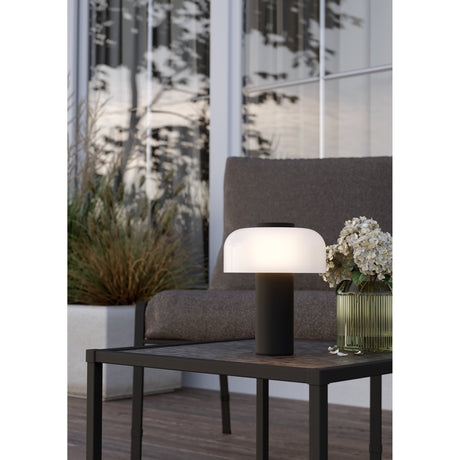 Ponente LED touch table lamp with black steel stem and frosted white glass dome, lit on an outdoor side table beside a sofa and vase of flowers.