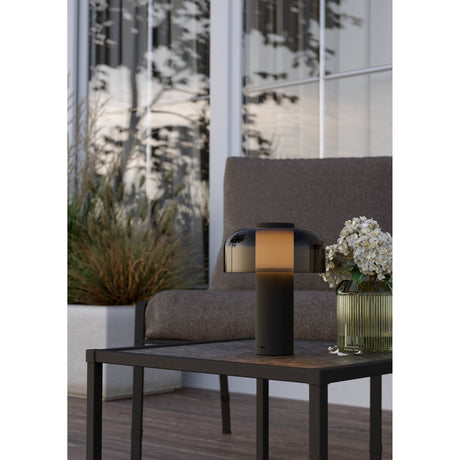 Ponente LED touch table lamp with black steel base and smoked mushroom-shaped dome shade, glowing warm on a side table beside a vase of flowers on an outdoor patio.