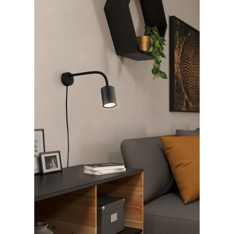 Portella Wall Light in black steel with round wall plate, slim curved arm and cylindrical spotlight head, shown illuminating a living room reading nook beside a sofa.