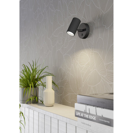 Portillo Wall Light in matt black aluminium and steel, with a fluted cylindrical head on a round backplate, shown illuminating a patterned wall above a shelf with books and a plant.