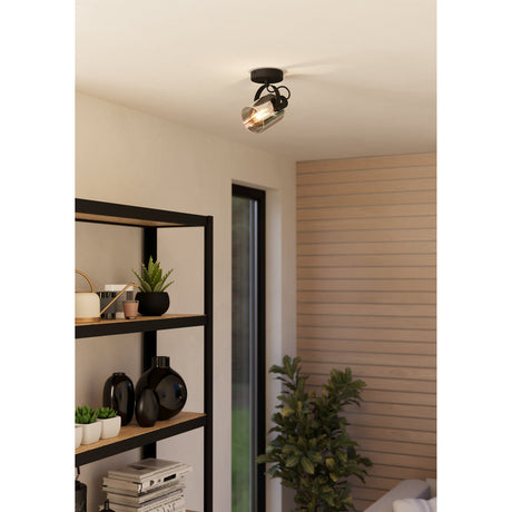 Povarne Wall Spotlight in black and chrome, single light with cylindrical smoked glass shade on a curved arm, mounted on a ceiling near a shelving unit in a living space.