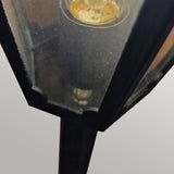 Close-up of the Parish 1 Light Pillar Light - Black, featuring a vintage-style geometric metal frame and glass panels, ideal for illuminating garden pathways. The light bulb inside emits a warm glow, adding outdoor lighting charm to the surrounding area. The background is a neutral, blurred grey.