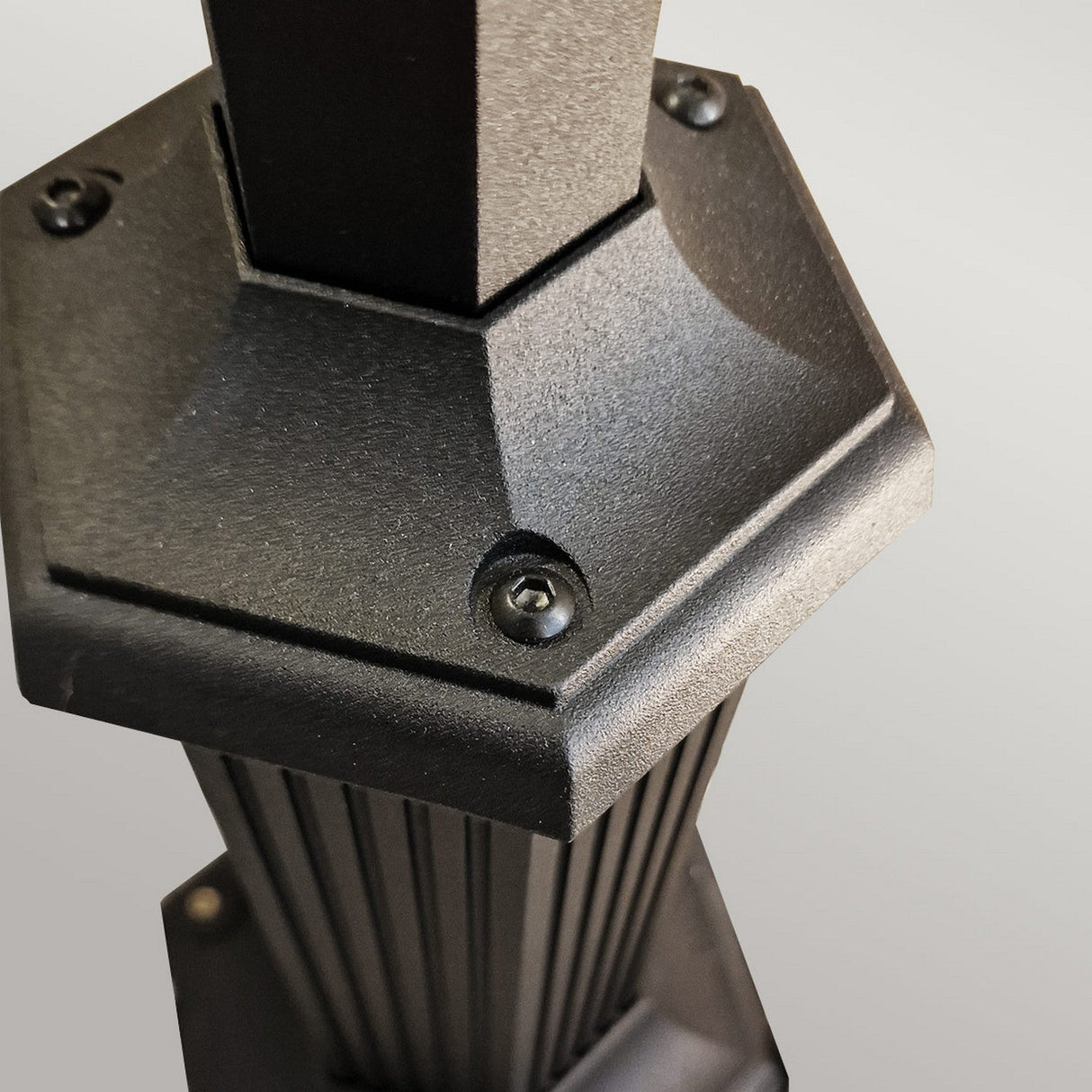 The image showcases a close-up of the Parish 1 Light Lamp Post in black, featuring a hexagonal metal structure made from die-cast aluminum. Visible screws and vertical grooves are evident on the slightly textured surface, with lighting accentuating the edges and details against a neutral backdrop, reminiscent of a sleek outdoor lamp post.