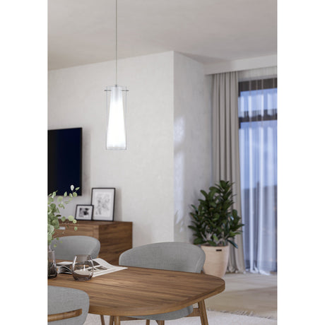 Pralbenne glass pendant light in chrome, with a clear outer cylinder and tapered white opal inner shade, hanging above a wooden dining table in a living room setting.