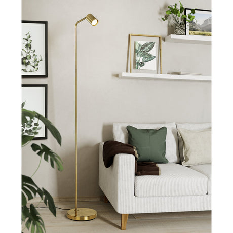 Pralmessa Floor Lamp in brushed brass with a slim steel stem and angled spotlight head, standing beside a sofa on a round base in a living room setting.