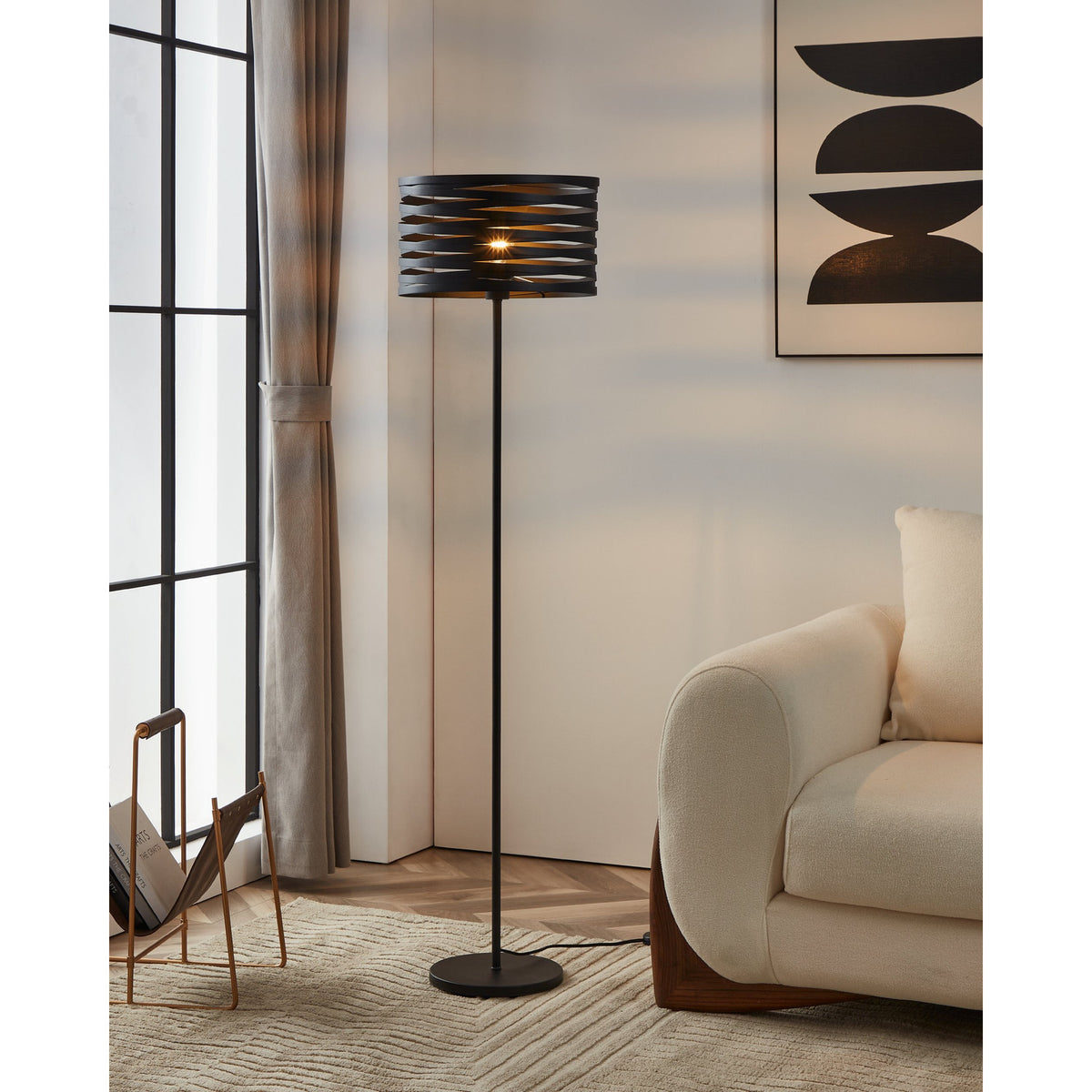 Pranvorne Floor Lamp in black steel with a slatted drum shade, slim stem and round base, shown lit beside a cream sofa in a living room.